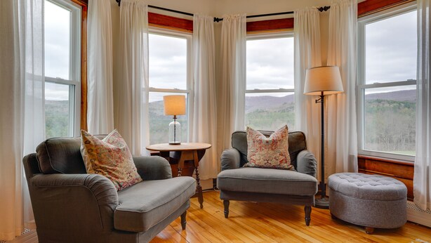 Une pièce chaleureuse avec deux fauteuils gris, un pouf assorti et une petite table avec une lampe. De grandes fenêtres, ornées de rideaux, offrent une vue sur l’extérieur.