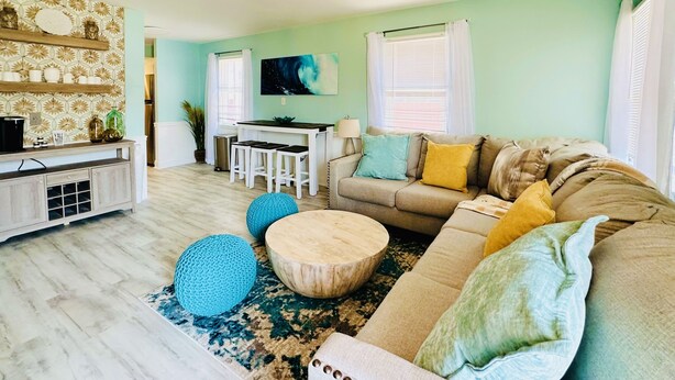 A living room with a beige sofa, round coffee table, and blue poufs. There is a dining area with white chairs and a table, and a kitchen area with a stove and cabinets.