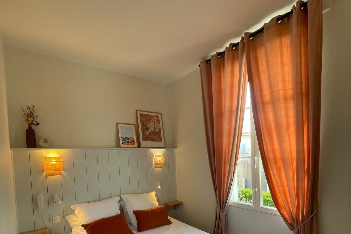A neatly made bed with white linens and orange pillows, a bedside table with a lamp, and a window with orange curtains.