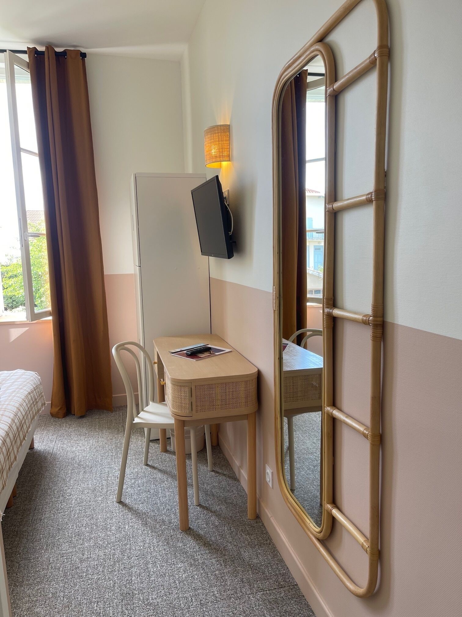 Une chambre d’hôtel comprenant un lit, un bureau avec une chaise, un téléviseur à écran plat fixé au mur, un grand miroir et une fenêtre avec des rideaux.