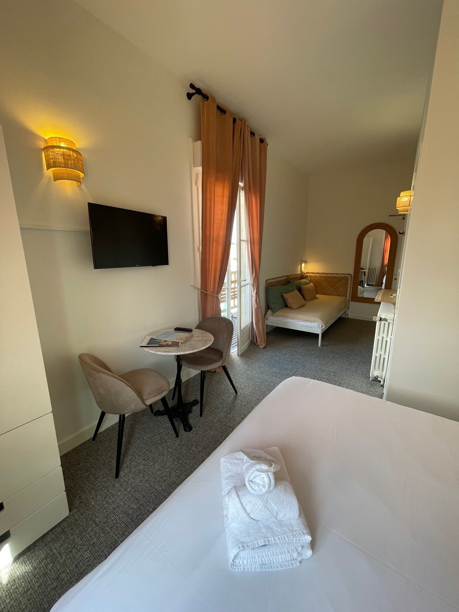 A hotel room with a bed, a desk with two chairs, a TV, a window with curtains, a sofa, and a mirror.