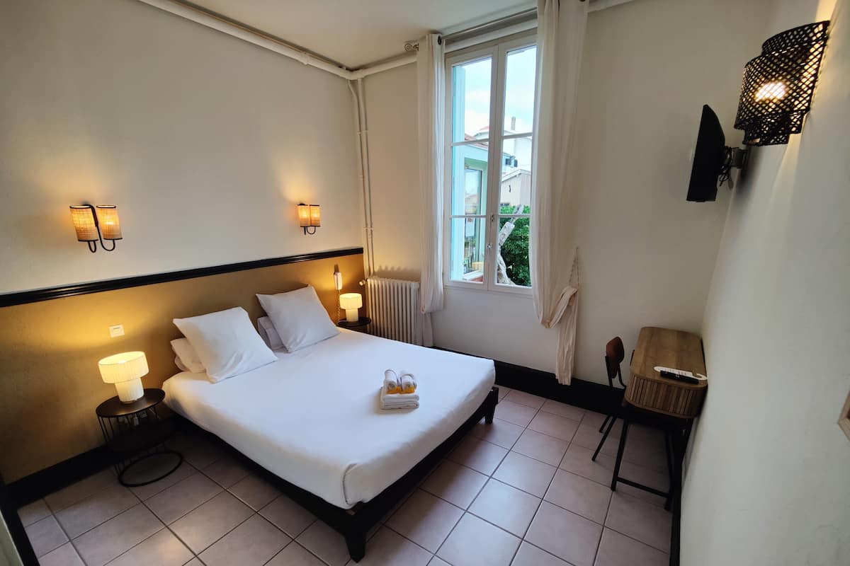 A hotel room with a large bed, bedside lamps, a window with curtains, a small wooden table, and a wall-mounted TV.