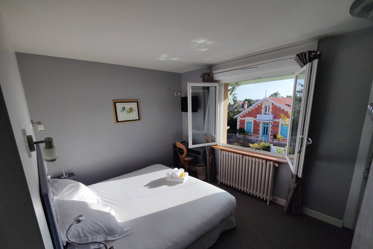 A hotel room with a bed, a window with a view of a building, a radiator, and a framed picture on the wall.