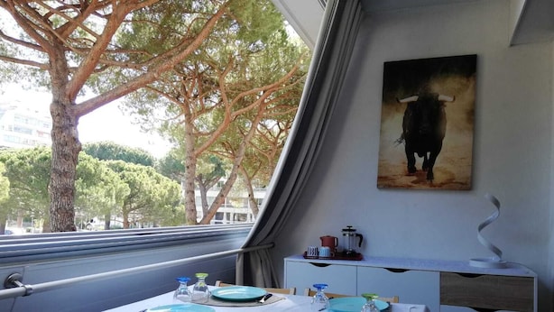 Un comedor con una mesa dispuesta para dos personas, un cuadro en la pared y vistas a árboles y edificios.