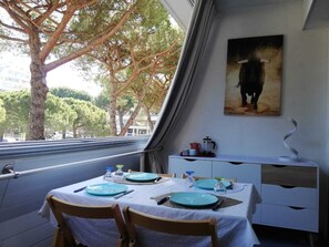 A dining area with a table set for two, a painting on the wall, and a view of trees and buildings outside.