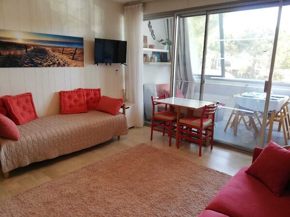 A cozy living room with a sofa, a dining table with chairs, a flat-screen TV mounted on the wall, and a large window offering a view of the outdoors.