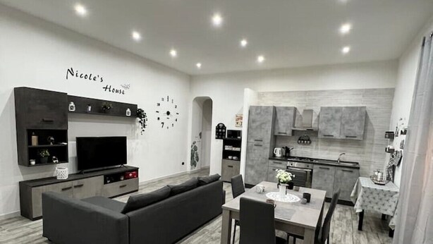 Ein modernes Wohnzimmer mit einem grauen Sofa, einem Esstisch mit Stühlen und einer Küchenzeile mit grauen Schränken.