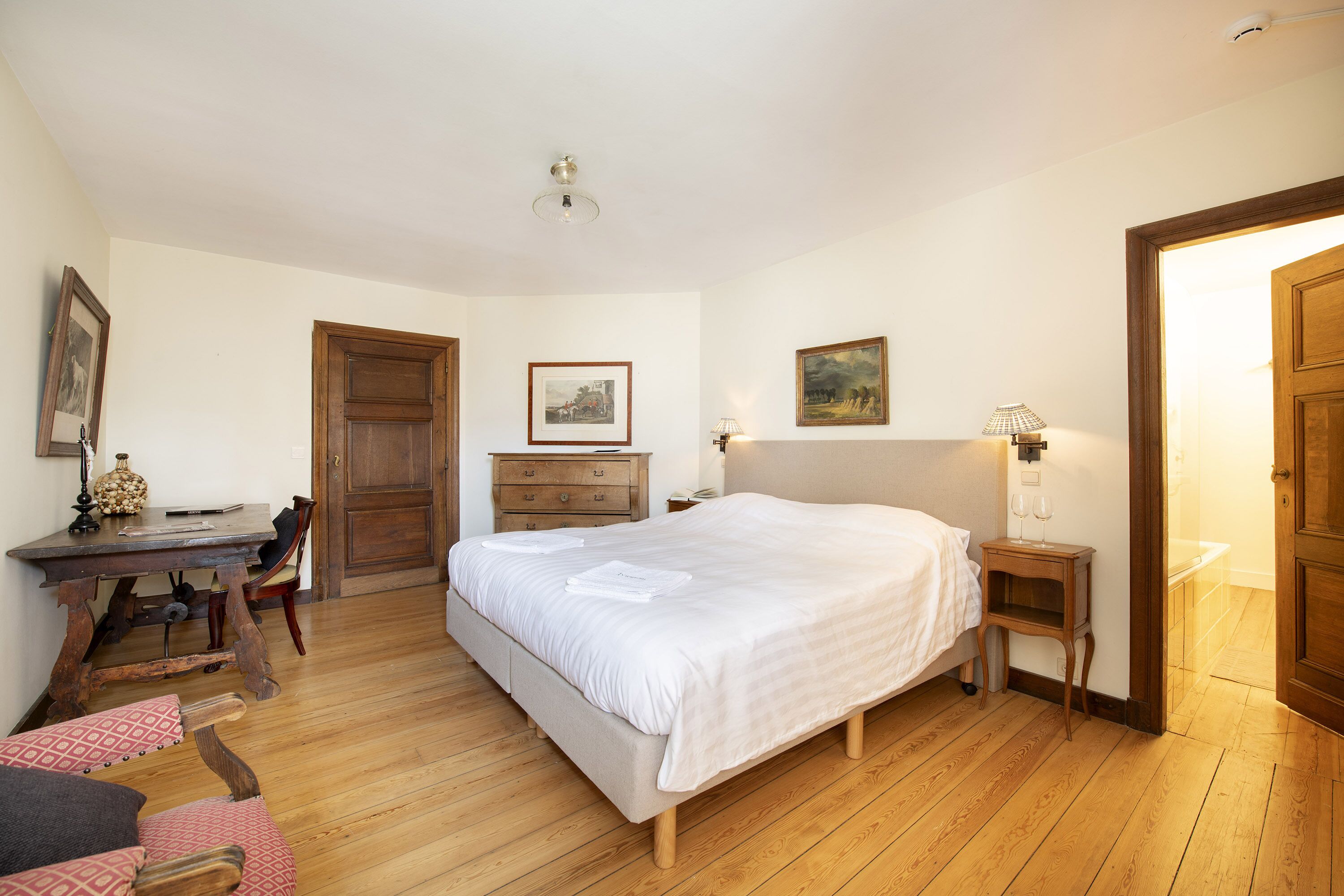 A bedroom with a large bed, wooden floor, a desk with a chair, a dresser, and two framed pictures on the wall.