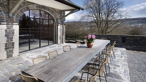 Eine Terrasse im Freien mit einem langen Holztisch, Korbstühlen, Steinmauern und Blick auf die Landschaft.
