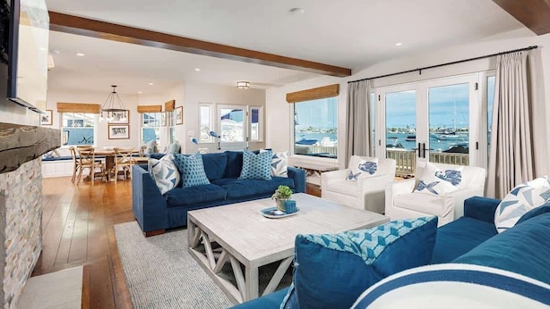 A spacious living area with a large sofa set, a coffee table, and a dining table with chairs. There are large windows offering a view of the sea and boats.