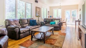 A spacious living room with leather sofas, a wooden coffee table, and large windows offering a view of the outdoors. - Magical Private House in Forest w/Hot Tub. Near Wineries! (Woodinville)