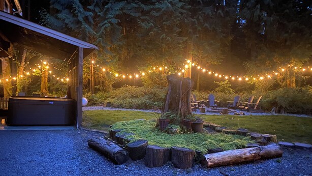 A cozy outdoor seating area with string lights, a fire pit, and a rustic wooden table. - Magical Private House in Forest w/Hot Tub. Near Wineries! (Woodinville)