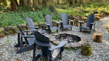 A fire pit surrounded by Adirondack chairs in a forested area with a gravel ground.