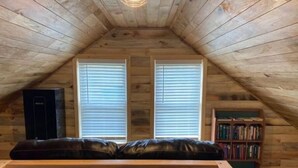 A cozy attic room with a sloped ceiling, two windows with blinds, a black leather sofa, a bookshelf, and a radiator. - Forest Side Solar Powered Cabin with Nature Trails (Norfolk)
