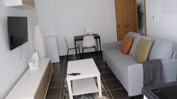 Un salon moderne avec un canapé gris, une table basse blanche, un téléviseur fixé au mur et une petite table avec des chaises.