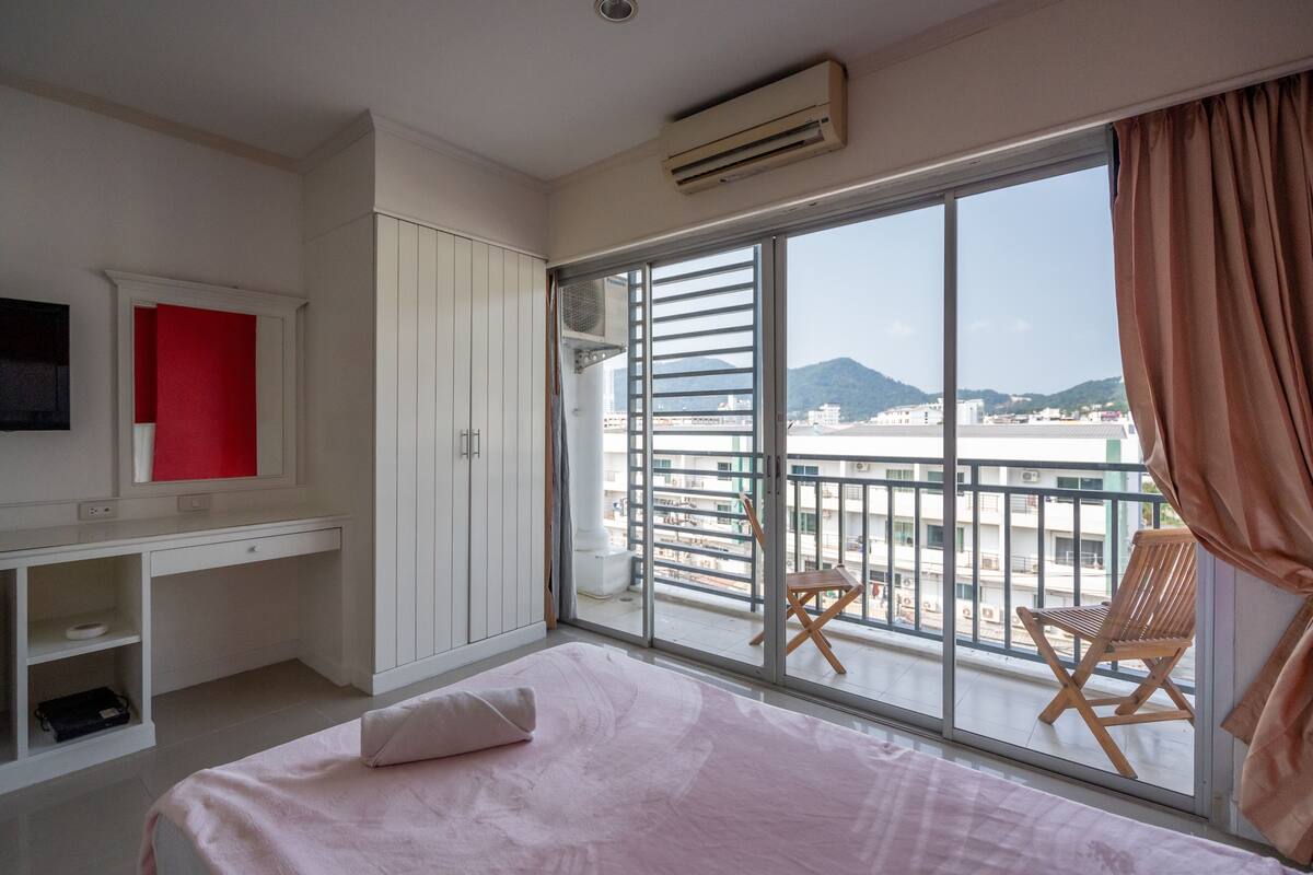 A bedroom with a bed, a desk, a chair, and a large window with a view of buildings and mountains.