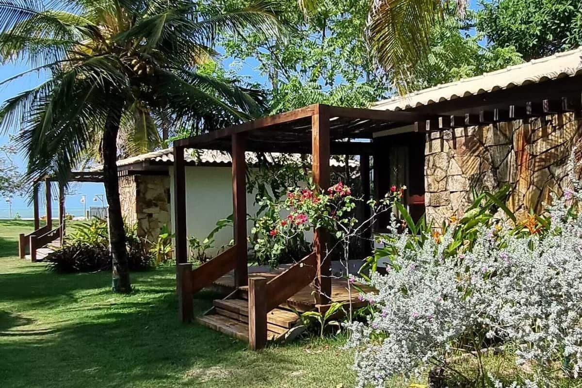 Ein tropischer Garten mit Palmen, einer Holzpergola und einem Steinhaus mit einer Treppe zu einem überdachten Bereich.