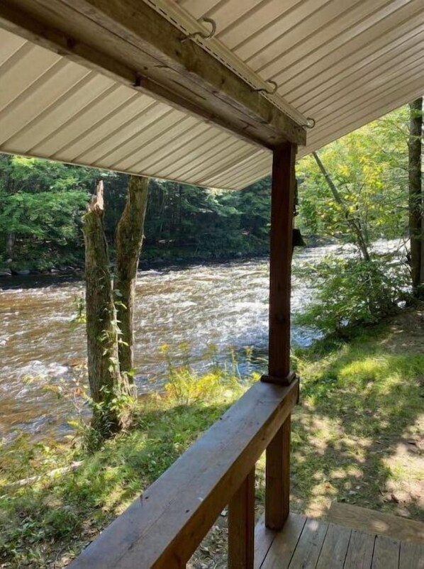 A wooden deck overlooking a river with trees and greenery in the background. - Riverside cabin, fly-fish for trout or just relax! (Malone)