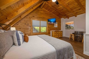 Cozy attic retreat with a large bed, wooden dresser, and TV, offering a peaceful view of the surrounding greenery.