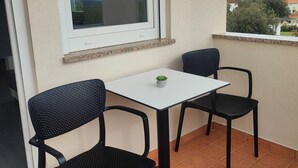 Un piccolo balcone con due sedie nere e un tavolo bianco, una finestra che si affaccia su un paesaggio verde, e un pavimento piastrellato.