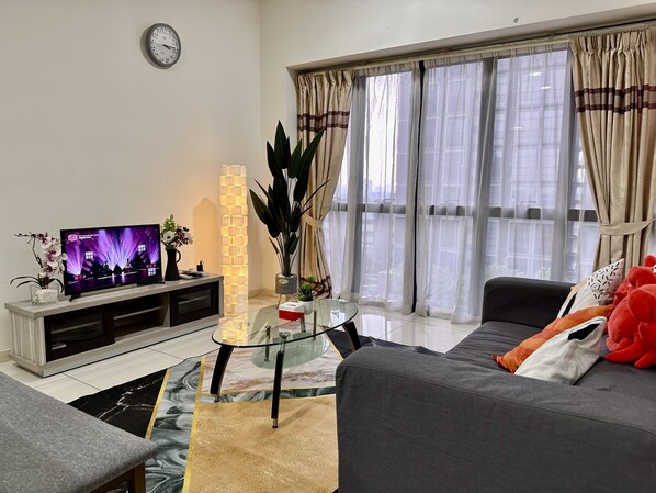 A living room with a flat-screen TV, a sofa, a glass coffee table, and a large window with curtains.