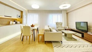 A modern living room with a sofa, chairs, a coffee table, a TV, and a kitchen area with cabinets and a microwave.