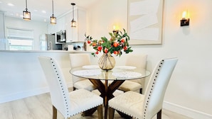 A dining area with a round glass table, four upholstered chairs, a vase with flowers, pendant lights, and a kitchen in the background. - Waterfront Condo, 8 min drive to Marco Beaches (Naples)