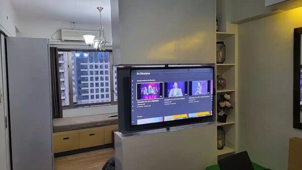 Un salon moderne avec un téléviseur à écran plat fixé au mur, un banc intégré le long de la fenêtre et une petite étagère ornée d’objets décoratifs.