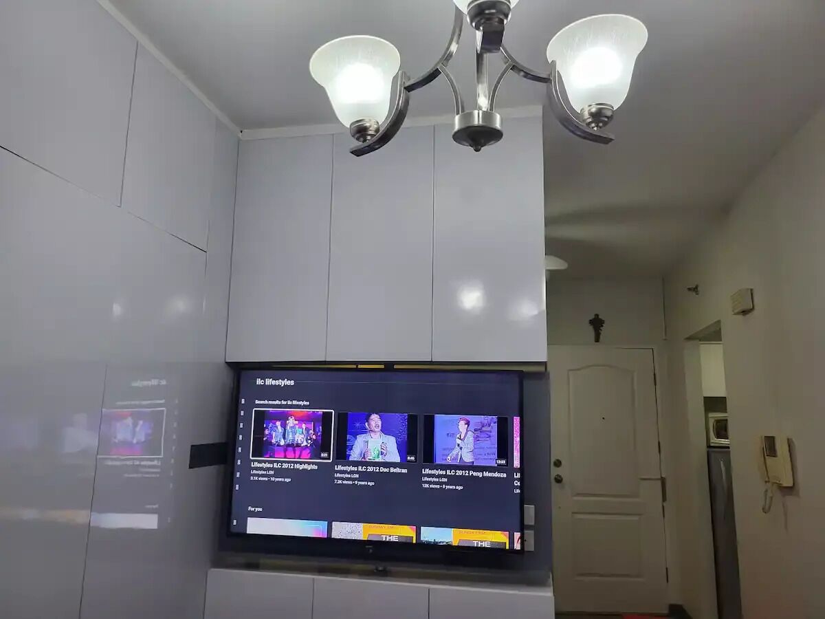 A room with a flat-screen TV mounted on a wall, a ceiling light fixture, and a door in the background.