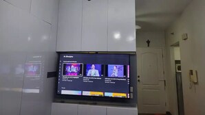 Une pièce avec un téléviseur à écran plat fixé au mur, un luminaire au plafond et une porte visible au fond.
