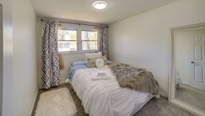Een slaapkamer met een bed, een raam met patroon gordijnen, een witte vloerkleed en een spiegel.