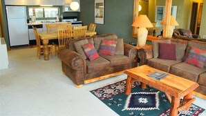 Un salon chaleureux avec deux canapés en cuir marron, une table basse en bois et un tapis à motifs. On aperçoit à l’arrière-plan un coin repas avec des chaises et une table en bois.