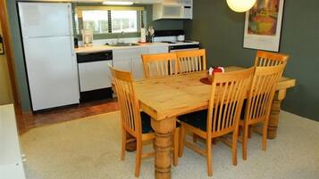 A compact kitchen with white appliances, a wooden dining table with six chairs, and a green wall with a framed picture.