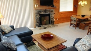 Een woonkamer met een stenen openhaard, een aan de muur bevestigde televisie, een eethoek met een tafel en stoelen, en een kroonluchter.