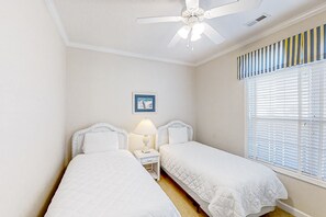 Un dormitorio con dos camas individuales, un ventilador de techo, una mesita de noche con lámpara, un cuadro en la pared y una ventana con persianas.