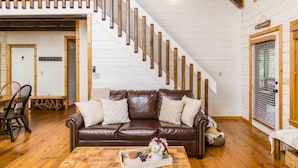 Een ruime woonkamer met een bruine leren bank, houten vloeren, een houten trap en een eethoek met een tafel en stoelen.