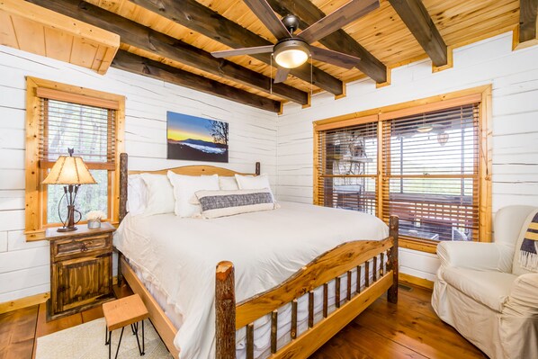 Una cama con estructura de madera, ropa de cama blanca, una mesita de noche con lámpara y una ventana con persianas.