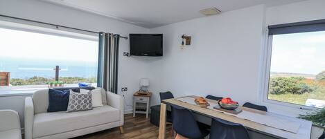 A modern living room with a sofa, dining table, and chairs, a flat-screen TV mounted on the wall, and a large window offering a view of the sea.