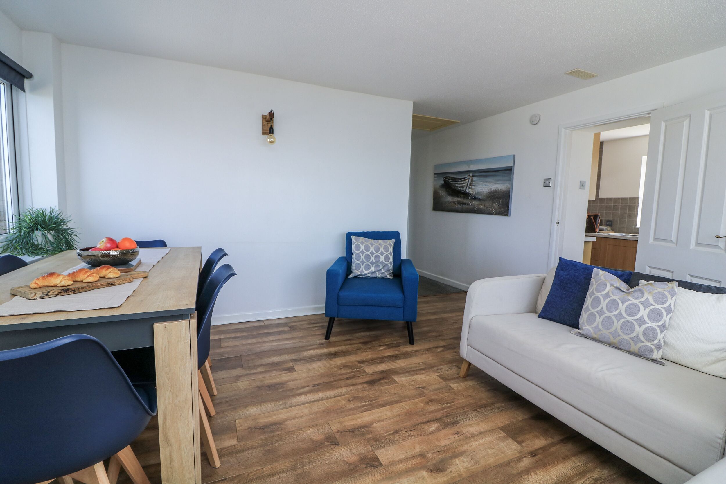 Ein Essbereich mit einem Holztisch, blauen Stühlen und einer Obstschale. Ein blauer Sessel und ein weißes Sofa mit gemusterten Kissen sind ebenfalls vorhanden. An der Wand hängt ein Gemälde und in der Ecke steht eine Pflanze.