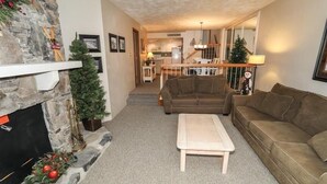 A cozy living room with a stone fireplace, a sofa, a coffee table, and a staircase leading to a kitchen. - Seven Springs 2 BR Deluxe Condo with Mountain Views by RedAwning (Champion)