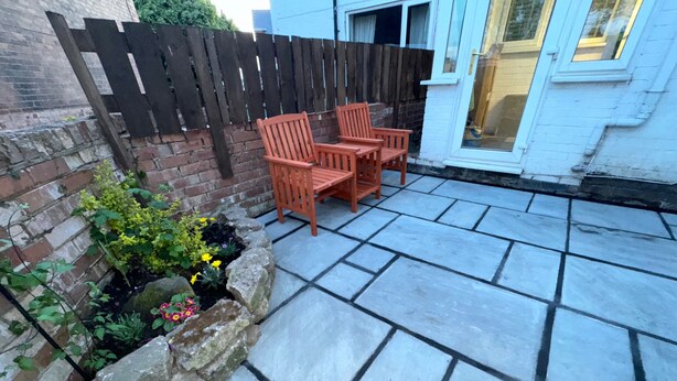 Une terrasse carrelée, une clôture en bois et un parterre agrémenté de plantes et de fleurs.