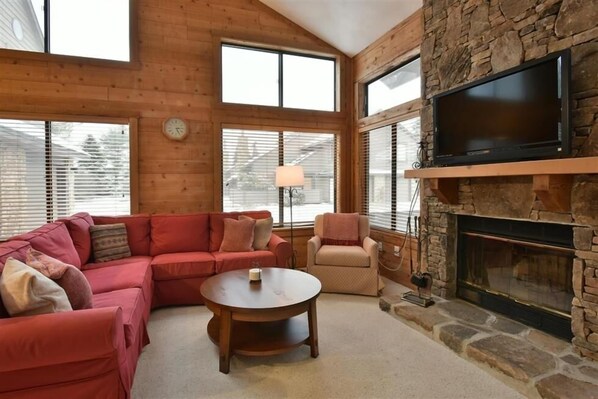 A cozy living room with a stone fireplace, a red sectional sofa, a round coffee table, and a television mounted on the wall. - Seven Springs Sunridge 3 BR Condo, Sleeps 10! by RedAwning (Champion)