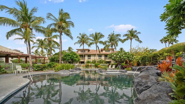 A pool surrounded by rocks and palm trees, with a building in the background.
