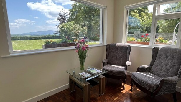 A room with two armchairs, a glass coffee table, and a vase of flowers. Large windows offer a view of a garden and distant hills. - Beautiful Irish Farmhouse an Hours Drive From Four Irish Cities (Clonmel)