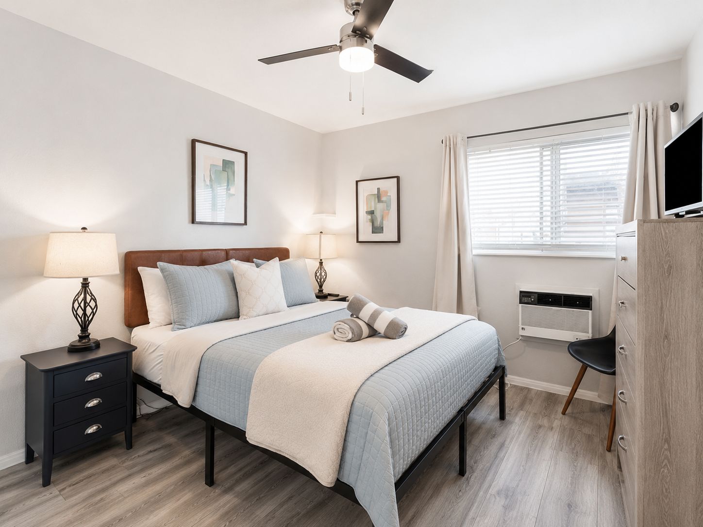 A bedroom with a bed, bedside tables, a ceiling fan, a window with curtains, and artwork on the wall.