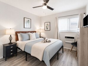 Een slaapkamer met een bed, nachtkastjes, een plafondventilator, een raam met gordijnen en kunst aan de muur.