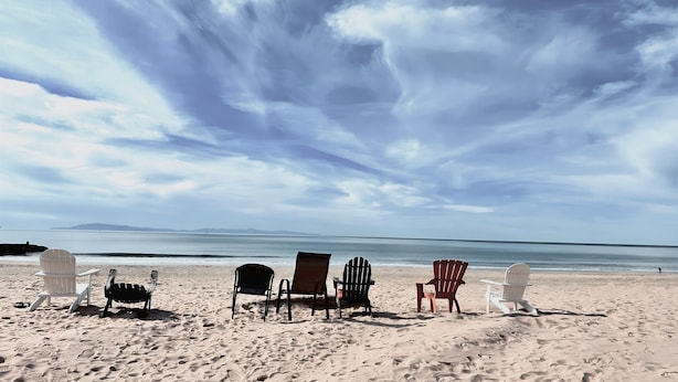 Une plage avec plusieurs chaises Adirondack et une table, donnant sur une mer calme sous un ciel nuageux.