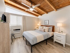 Une chambre à coucher avec un lit, des lampes de chevet, un ventilateur de plafond, une fenêtre avec des rideaux, un radiateur et un téléviseur posé sur un meuble.