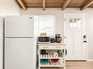 Cocina compacta con refrigerador blanco, microondas, cafetera y un mueble pequeño con utensilios de cocina y ensaladeras.