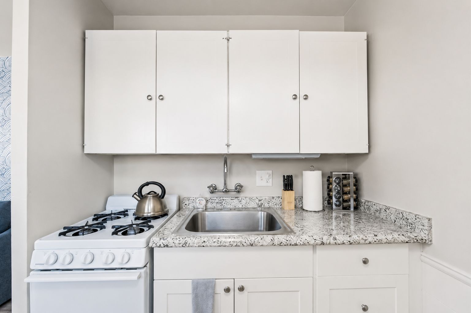 Une cuisine compacte avec des meubles blancs, un évier en acier inoxydable, une cuisinière à gaz et une bouilloire.